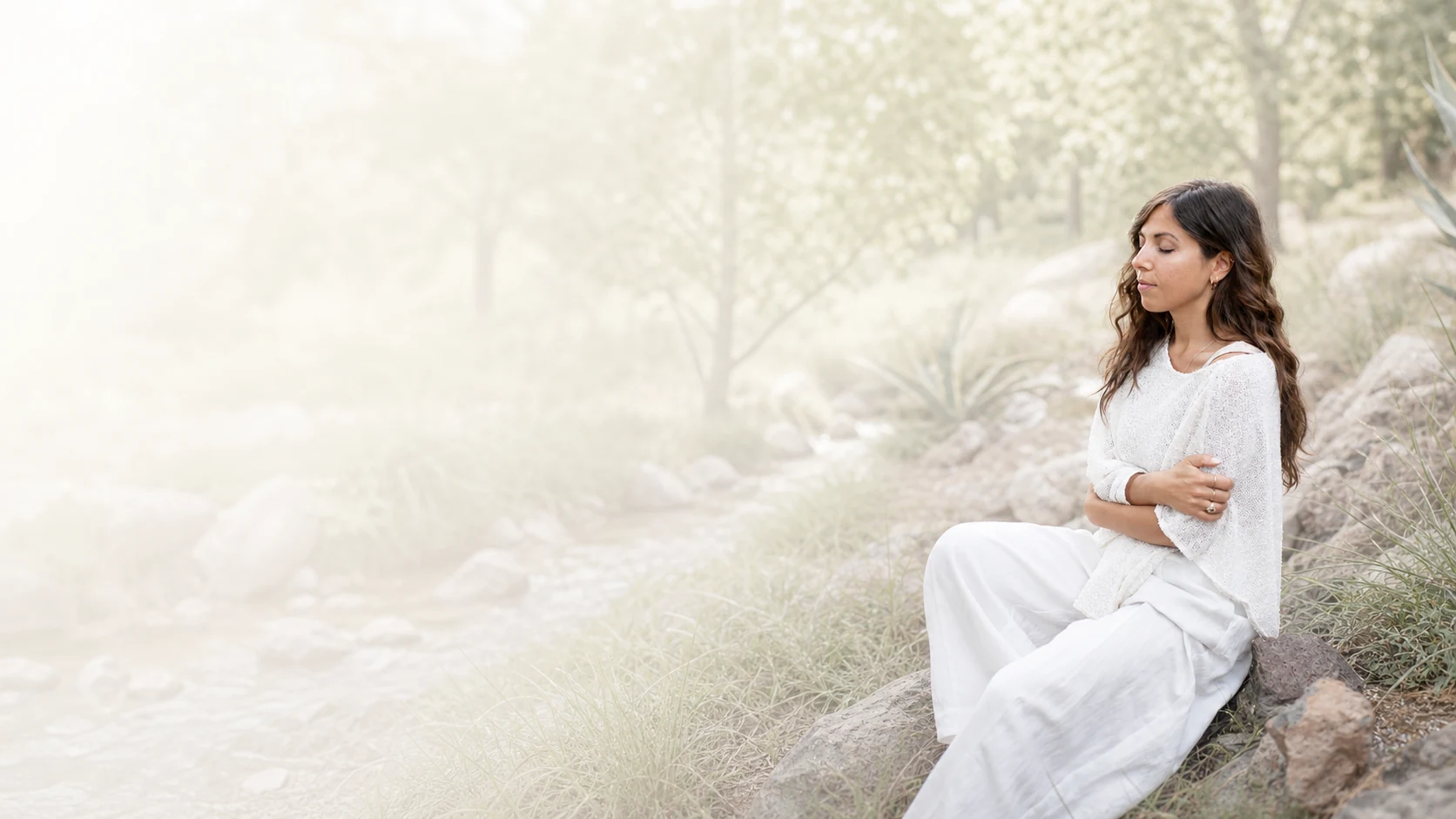 Manuela seated outdoors, gently embracing herself in a calm reflective gesture.